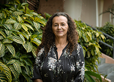 Fotografia mostra, em primeiro plano, a professora Ana Cleide Moreira vestindo camisa preta com estampas brancas. Ela tem cabelos encaracolados abaixo dos ombros e sorri para nós. No fundo, folhagem natural verde e amarela.
